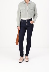 Giacca in velluto a coste verde chiaro cropped con due tasche frontali, abbinata a jeans a vita alta blu scuro e ballerine beige, mentre tiene una borsa marrone.