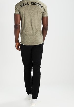 Uomo in piedi con le spalle rivolte allo spettatore, che indossa una t-shirt grigio chiaro con testo "HELL RIDERS", pantaloni neri e sneakers bianche.