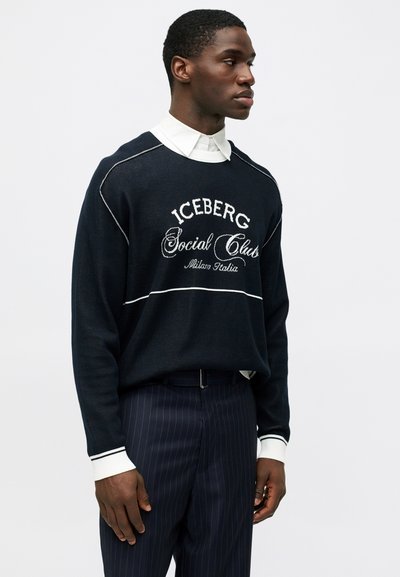 Άνδρας που φοράει ναυτικό πουλόβερ με λευκό κείμενο "ICEBERG Social Club Milano Italia", λευκό πουκάμισο και ναυτικές ραβδωτές παντελόνες, στραμμένος προς τα δεξιά.