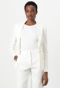 Weißer maßgeschneiderter Blazer über einem weißen Hemd, kombiniert mit passenden, hochtaillierten Hosen. Glatter Stoff, strukturiertes Design, minimalistischer Stil.