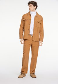 Bruine corduroy shirtjack met twee borstzakken, gecombineerd met bijpassende broeken. Gekozen voor een witte coltrui en beige schoenen.