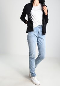 Svart zip-up jacka med huva, grå t-shirt, ljusblå skinny jeans och vita sneakers. Jackan har sidofickor och en slät yta.