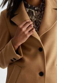 Mantel aus Kamelwollmischung mit Reverskragen, schwarzen Knöpfen und Seitentaschen, darüber getragen eine gemusterte Bluse. Glatte Textur.