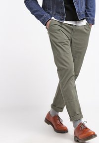 Person som bär olivgröna byxor, grå strumpor, bruna läderskor, en svart tröja, vit skjorta och en blå denimjacka med händerna i fickorna.