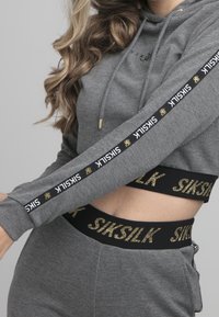 Grijze cropped hoodie met lange mouwen, voorzien van zwarte en witte merktekens. Zwarte tailleband met gouden tekstaccenten. Zachte textuur.
