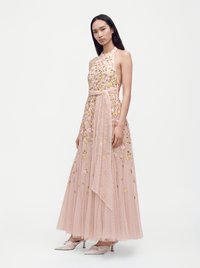 Femme portant une robe rose sans manches brodée de fleurs avec une ceinture transparente et des chaussures à talons bas pointues, debout sur un fond blanc.
