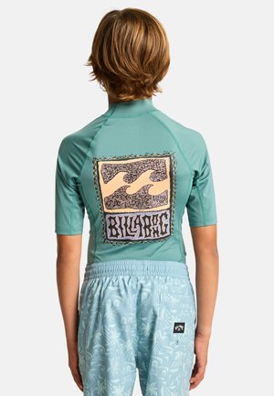 Bambino che indossa una maglietta da bagno a maniche corte color verde acqua con il logo Billabong e pantaloncini da bagno azzurri con motivo, visto di schiena.