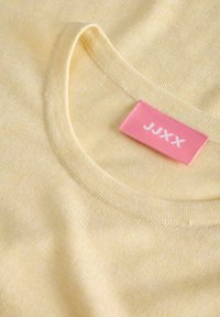 Gros plan sur un pull ras du cou jaune pâle montrant un tissu en tricot doux et une étiquette de vêtement rose avec "JJXX" imprimé en blanc.
