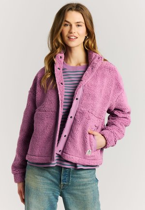 Jonge vrouw die een roze sherpa fleece jas draagt over een paars-groen gestreept shirt met blauwe spijkerbroek, poserend met één hand in de jaszak.