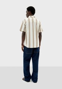 Camisa de manga corta en crema con rayas verticales marrones, con cuello inglés y dobladillo recto, combinada con pantalones de mezclilla azul oscuro.
