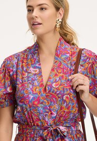 Színes virágmintás ruha rövid ujjakkal, mély V-kivágással és derékra köthető övvel. Rózsaszín, lila és narancssárga árnyalatok jellemzik. Arany fülbevaló és barna táska.