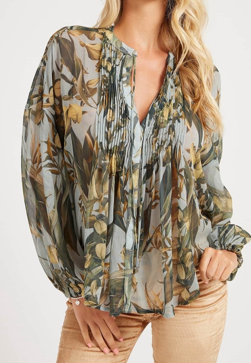 Femme portant un chemisier transparent à imprimé floral avec des détails plissés et des manches longues bouffantes, assorti à un pantalon en velours côtelé marron clair.