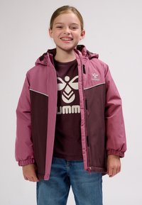 Smilende barn iført en todelt jakke i pink og bordeaux over en bordeaux skjorte med en stor logo og blå jeans, stående mod en ensfarvet baggrund.