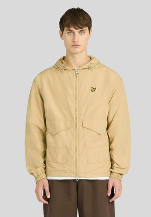 Ung mand iført en beige hættejakke med lynlås, klaplommer og et lille gult fugleemblem på brystet mod en ensfarvet baggrund.
