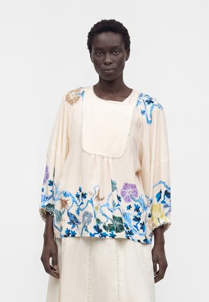 Femme portant un chemisier ample crème avec une broderie florale bleue, violette et marron et des manches larges, assorti à une jupe crème.