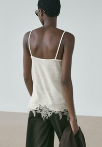 Frau mit kurzen lockigen Haaren, trägt ein weißes Top mit Spitzenbesatz und schwarze Hose, hält eine dunkle Jacke und steht mit dem Rücken zu einem schlichten Hintergrund.