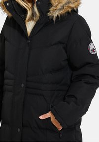 Femme portant une doudoune noire avec capuche bordée de fourrure, main dans la poche zippée, et écusson rond sur le haut de la manche.