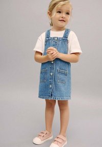 Džínová overal šaty se dvěma předními kapsami, světle modrá barva, kovové knoflíky, v kombinaci s bílým tričkem a růžovými nazouvacími botami.