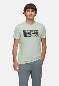 Ljusgrön T-shirt med en svart abstrakt grafik och texten "MAMMUT" tryckt på framsidan. Kortsleeves och rund halsringning.