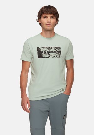 Lichtgroen T-shirt met een zwart abstract grafisch ontwerp en de tekst "MAMMUT" op de voorkant gedrukt. Korte mouwen en een ronde halslijn.