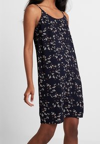 Žena v bezrukávových tmavě modrých šatech s tenkými ramínky a malým bílým a červeným květinovým vzorem, stojící na jednobarevném pozadí.