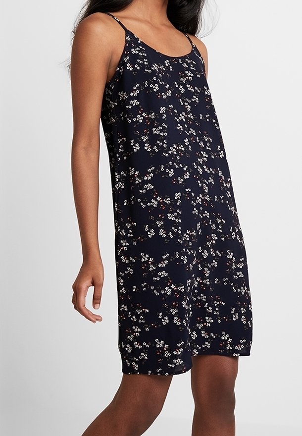 Žena v bezrukávových tmavě modrých šatech s tenkými ramínky a malým bílým a červeným květinovým vzorem, stojící na jednobarevném pozadí.