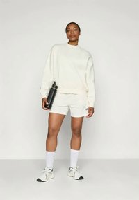 Hvit fleece hettegenser og matchende shorts, holdt på en rustfritt stål vannflaske, hvite treningssokker og sorte sko. Glatte teksturer, uformell design.