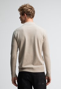 Beige Strickpullover mit geripptem Kragen und Bündchen. Glatte Textur, figurnah geschnitten und schlichtes Design. Über dunklen Hosen getragen.