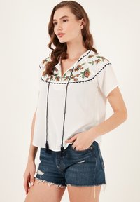 Valkoinen broderad blus med blommönster, marinblå kant och dragsnöre vid halsen. Kombineras med denimshorts med fransigt fåll.