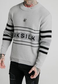 Man med grå, stickad tröja med svarta ränder och texten "SIK SILK", svarta slitna jeans samt synliga tatueringar på hals och händer.
