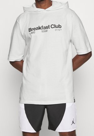 Mężczyzna ubrany w białą koszulkę z kapturem i krótkim rękawem z napisem „Breakfast Club” oraz czarno-białe sportowe spodenki z logo Jordana.