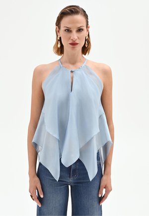 Femme portant un top à bretelles superposé bleu clair et un jean bleu, debout devant un fond blanc uni.