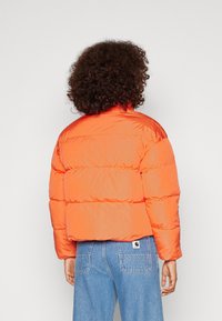 Giacca puffer arancione con design cropped, caratterizzata da una finitura lucida e sezioni trapuntate orizzontali. Indossata su jeans di denim blu.