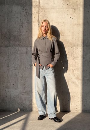 Une femme blonde pose devant un mur en béton, vêtue d’une chemise à carreaux ceinturée, d’un jean ample délavé clair et de chaussures noires.