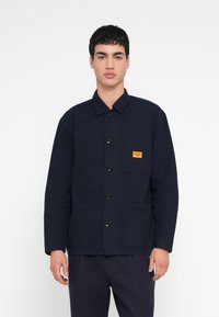 Μπουφάν από καμβά σε ναυτικό μπλε χρώμα με κλείσιμο με κουμπιά μπροστά, δύο πλαϊνές τσέπες, τσέπη στο στήθος με πορτοκαλί λογότυπο και γιακά.