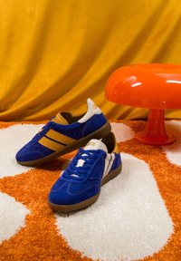 Paire de baskets bleues et beige sur un tapis à motifs orange et blanc à côté d'une petite table ronde orange sur un fond jaune.