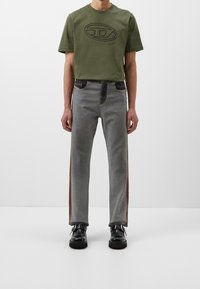 Camiseta verde oliva con logotipo en relieve, combinada con jeans grises claros que presentan bolsillos oscuros y franjas laterales marrones. Unos zapatos negros completan el conjunto.