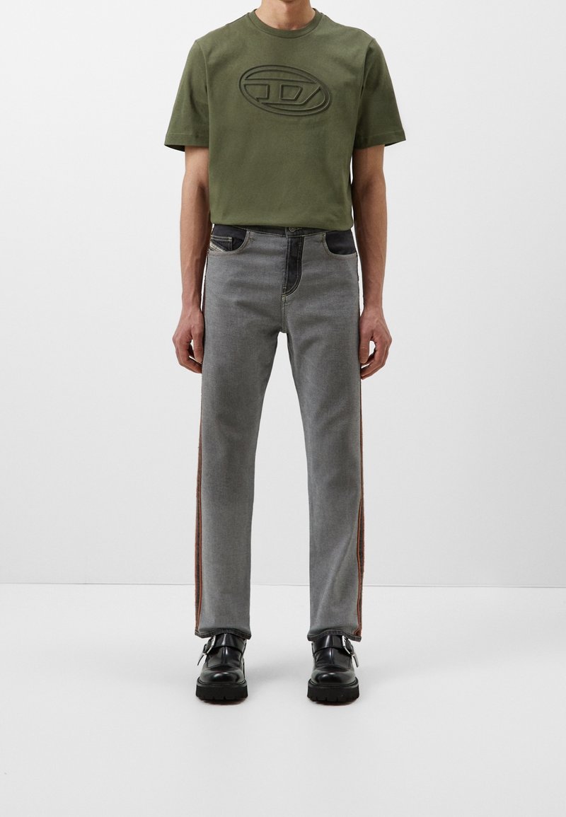 Camiseta verde oliva con logotipo en relieve, combinada con jeans grises claros que presentan bolsillos oscuros y franjas laterales marrones. Unos zapatos negros completan el conjunto.