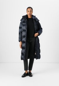 Doudoune bleu marine à la coupe longue et matelassée, avec des accents en métal doré, une capuche et des poches latérales, assortie à un jean noir et des mocassins.