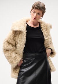 Beige faux fur jakke med tekstureret finish, bred krave og løs pasform, parret med en tætsiddende sort top og lædernederdel.