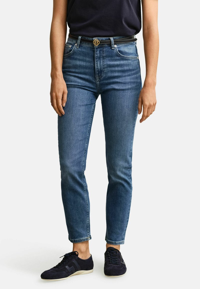 Osoba stojící proti jednobarevnému bílému pozadí, oblečená v modrých džínech se středním pasem, černým páskem a černých teniskách.