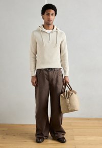 Sweatshirt com capuz em malha texturada creme clara, com fecho de correr, combinada com calças castanhas de perna larga. Segura uma mala em pele castanha com alças contrastantes.