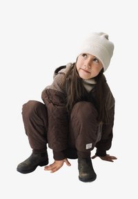 Veste matelassée marron et pantalon avec des accents en molleton, assortis à un bonnet en tricot côtelé crème et des bottes sombres, sur un enfant accroupi.