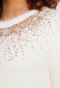 Valkoinen pörröinen neule, jossa on pyöreä pääntie, ja ylhäällä on epätasaisesti aseteltuja kultaisia ja hopeisia paljetti, luoden kimaltelevan efektin.