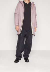 Homme portant une longue veste à capuche violet clair par-dessus un t-shirt noir, un jean noir ample avec des détails sur le panneau avant, et des chaussures noires à lacets.