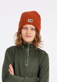 Gorro de punto acanalado naranja con un parche de logo en la parte frontal. Se lleva con una sudadera verde oscura con cremallera que tiene un logo a lo largo de la cremallera.
