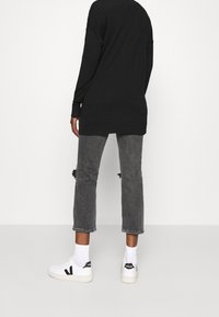 Persona vestida con una camiseta de manga larga negra, jeans cortos grises desgastados, calcetines blancos y zapatillas Veja blancas, de pie contra una pared lisa.
