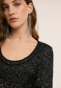 Czarny, teksturowany sweter z dzianiny z dekoltem w kształcie litery U, ozdobiony metalicznymi nitkami. Długie, srebrne kolczyki z frędzlami dodają charakteru.