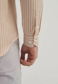 Chemise à manches longues rayée marron clair avec des rayures blanches, dotée de poignets à boutons et d'une texture lisse. Coupe ajustée visible à l'ourlet.