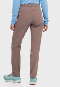 Braune, dehnbare Wanderhosen mit einem elastischen Bund in mittlerer Höhe, Seitentaschen und tapered Beinen, kombiniert mit einem blauen Longsleeve-Oberteil und leichter Fußbekleidung.